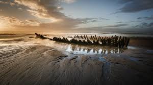 A wrecked boat sits on a sandy beach at sunset, with the sky filled with orange and grey clouds.