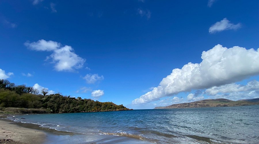 A coastal landscape features a sandy beach meeting a blue sea, with a green headland visible under a cloudy blue sky.