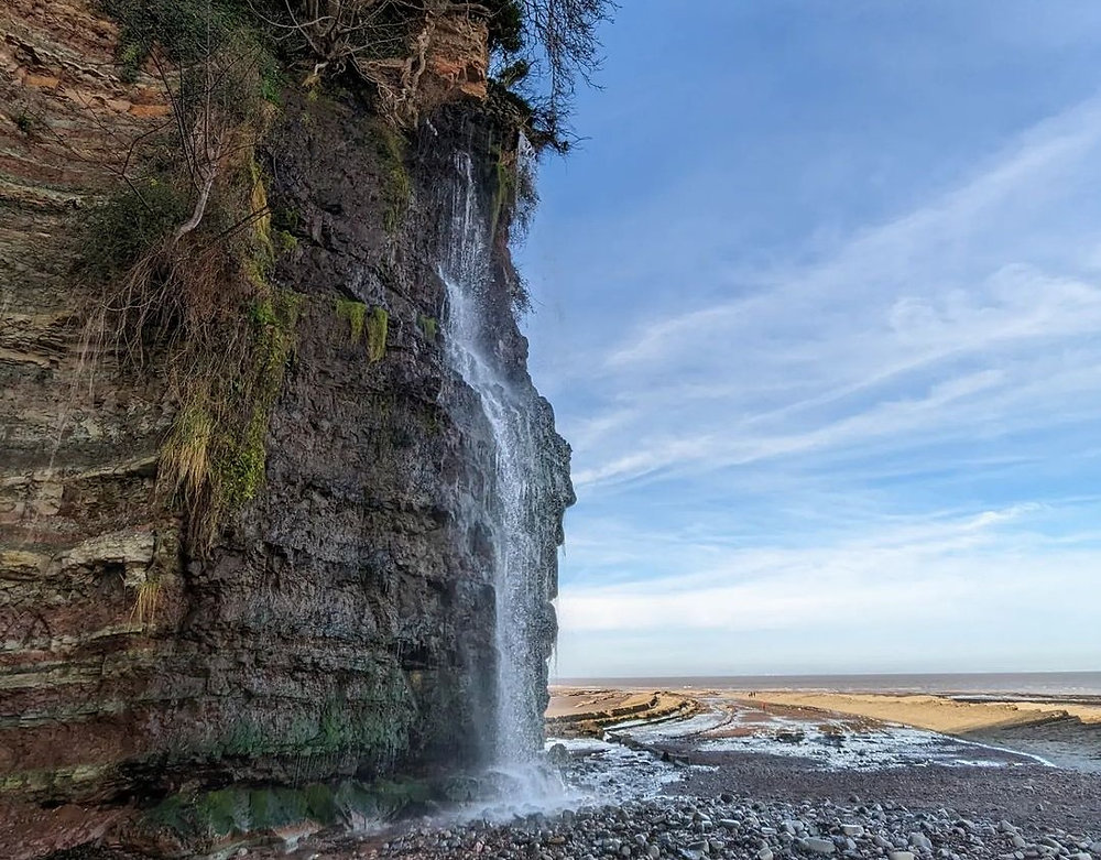 A waterfall cascades down a dark cliff onto a rocky beach, with the sea visible in the background under a cloudy sky. | St Audries Bay
