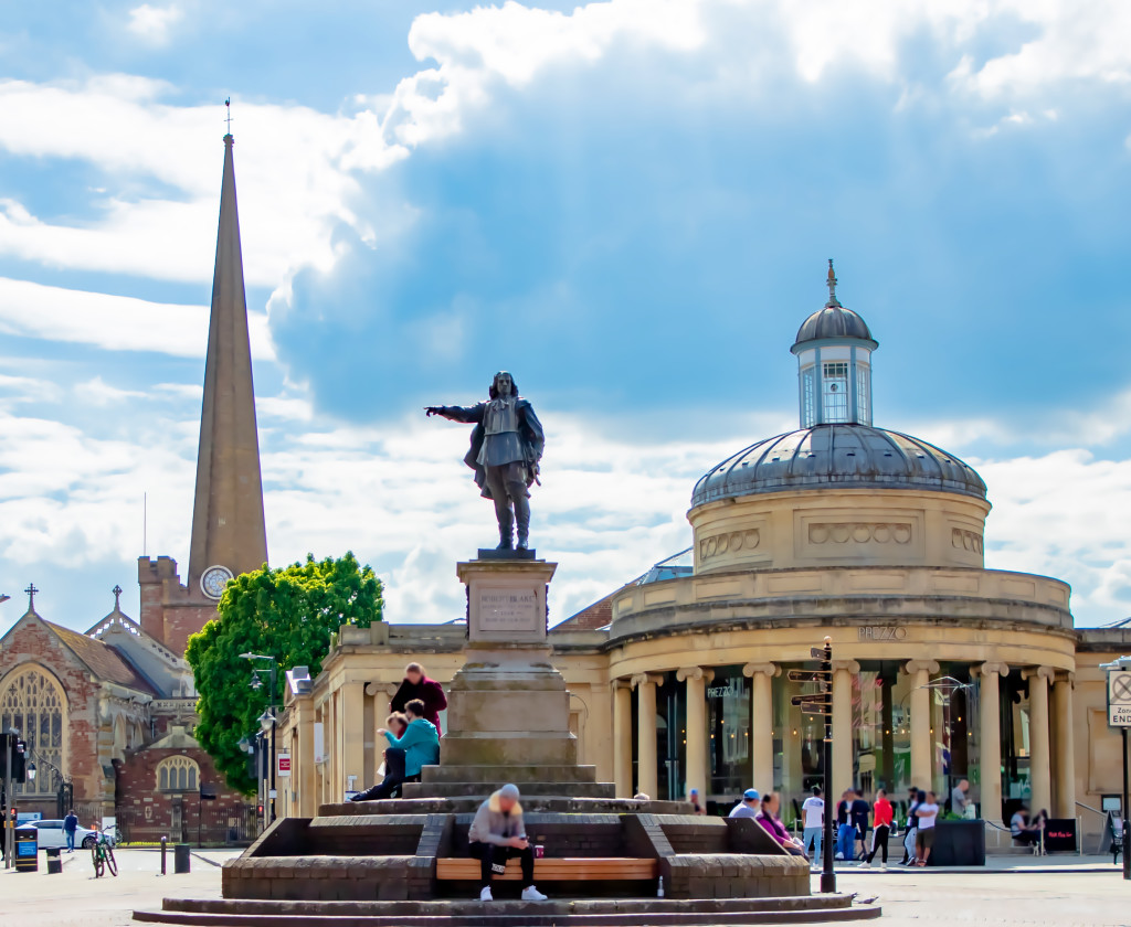 A statue of Robert Blake stands on a stone pedestal in Bridgwater. Behind the statue, there is a building with a domed roof and St Mary's Church tall spire. People are walking and sitting around the square.