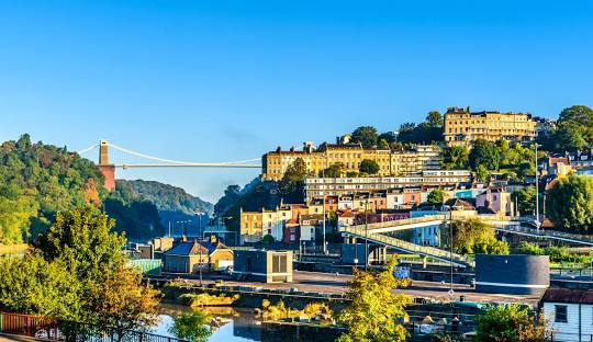 Clifton Suspension Bridge spans the Avon Gorge in Bristol, with colourful buildings lining the hills under a clear blue sky.
