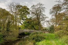A stone bridge over a river with a grassy path to the right and tall trees in the distance, Part of the Coleridge Way.