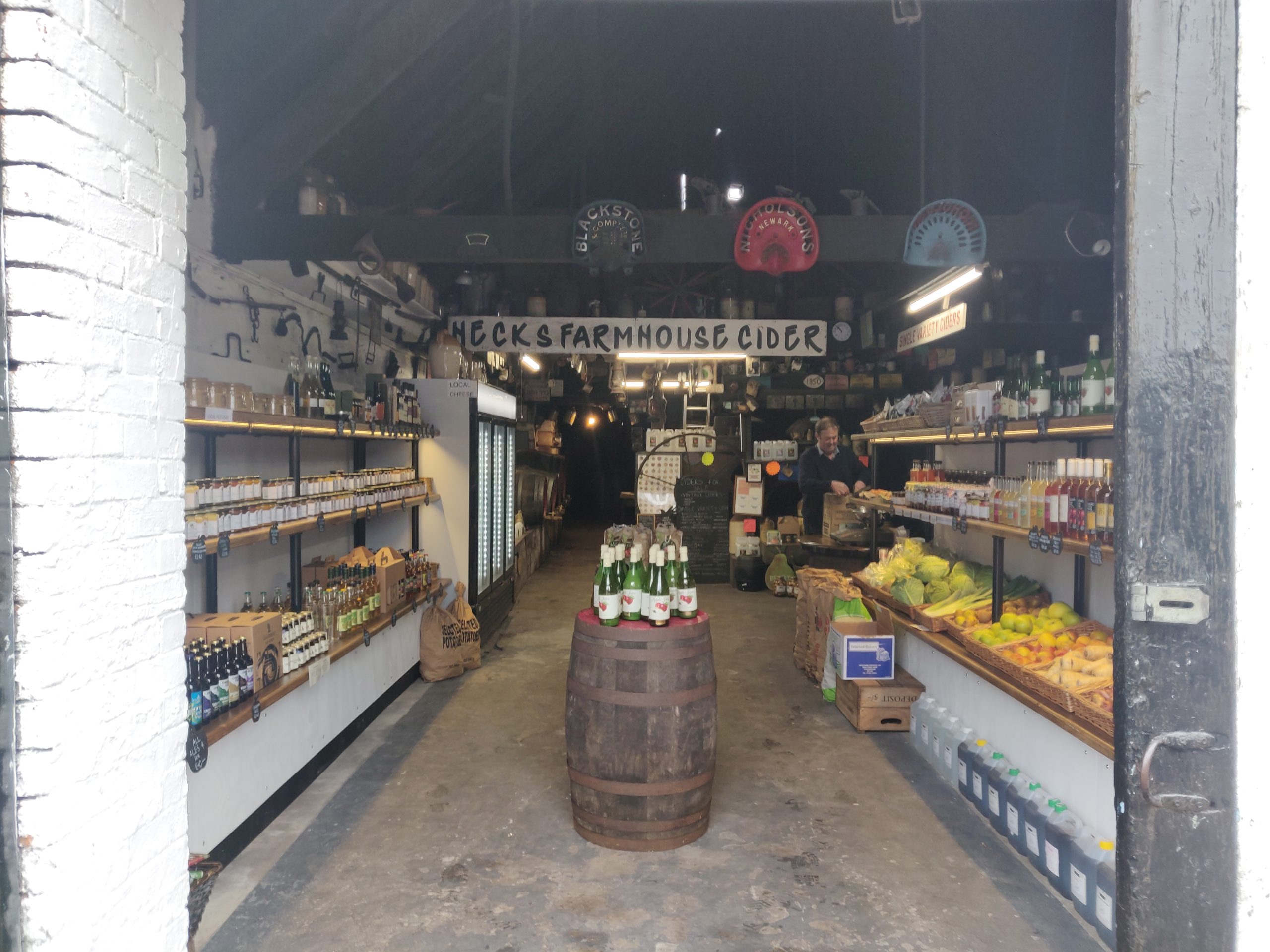 The entrance to Hecks Farmhouse Cider shop | In the centre is a barrel topped with bottles. Either side of that are shelves filled with fresh produce.