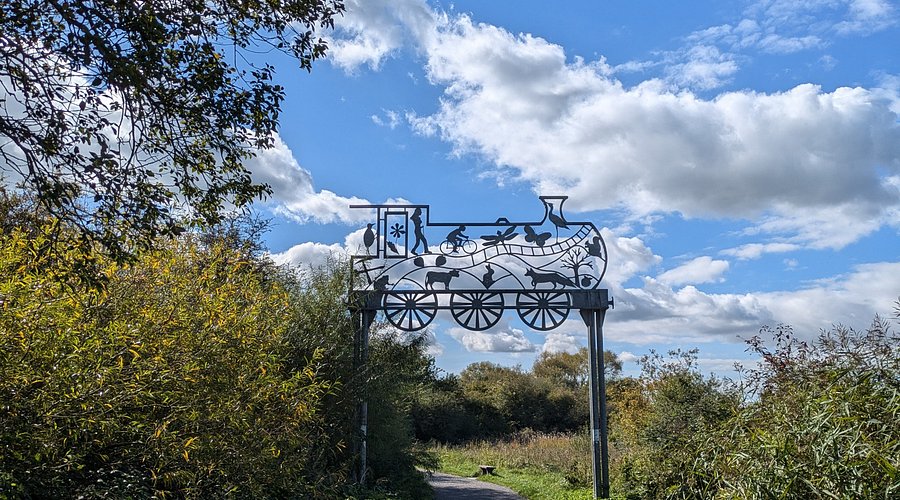 A metal open work sign depicting animals, people and wildlife within the shape of a steam train above the path of the Strawberry Line.