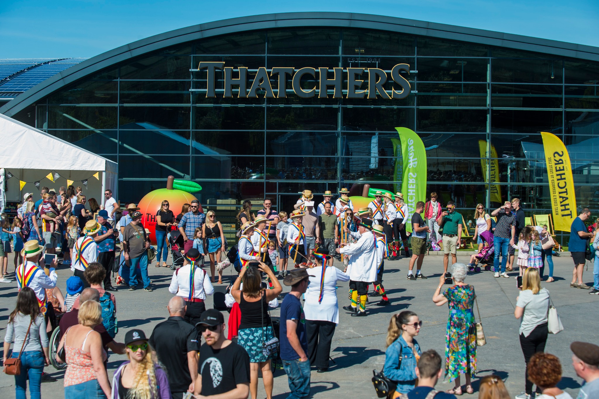 Large glass building with Thatchers written on the front. Outside there is a group of Morris Dancers and many spectators