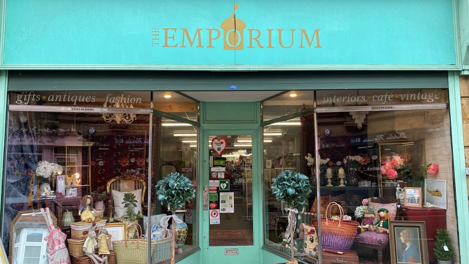A shop front with a light blue sign with The Emporium written in gold. there is an eclectic mix of bags, mirrors, pictures and furniture displayed in the windows.
