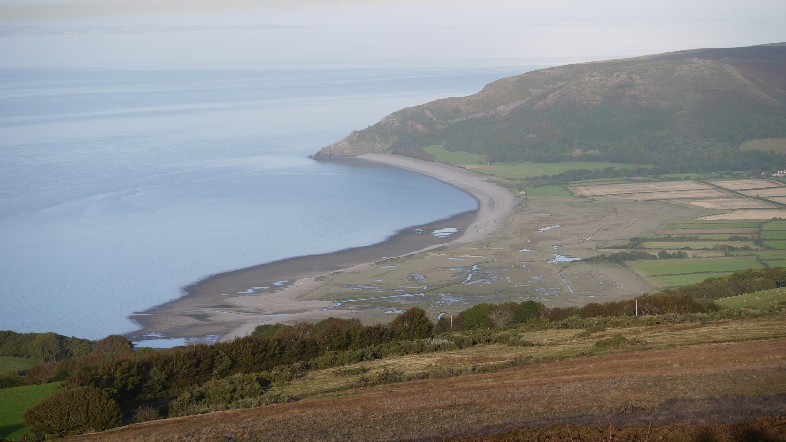 A curved bay with blue sea to the left and fields and hills to the right as views from Exmoor | Southwest Coastal Path