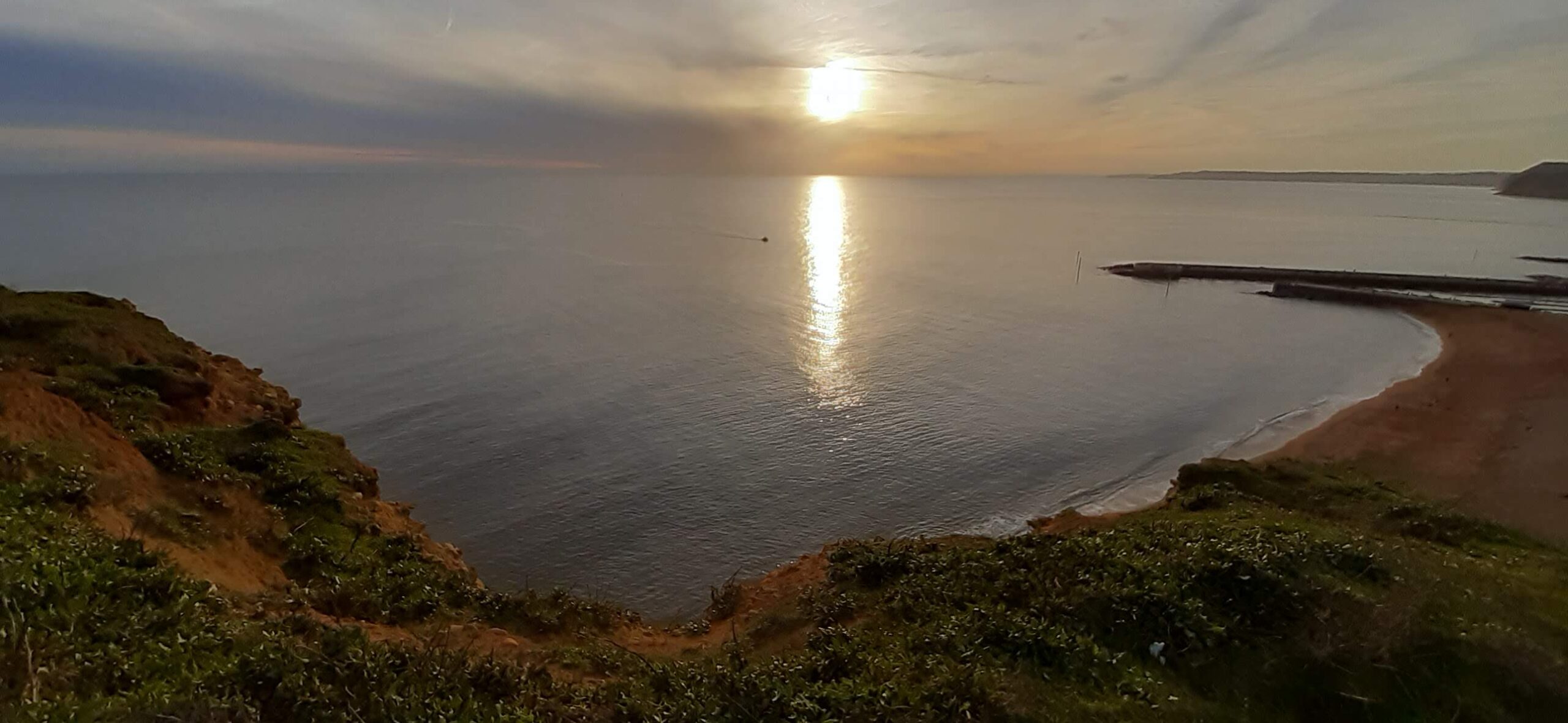 A coastal view shows a cliff overlooking the sea at sunset, with the sky filled with orange and grey clouds. | Jurassic Coastline